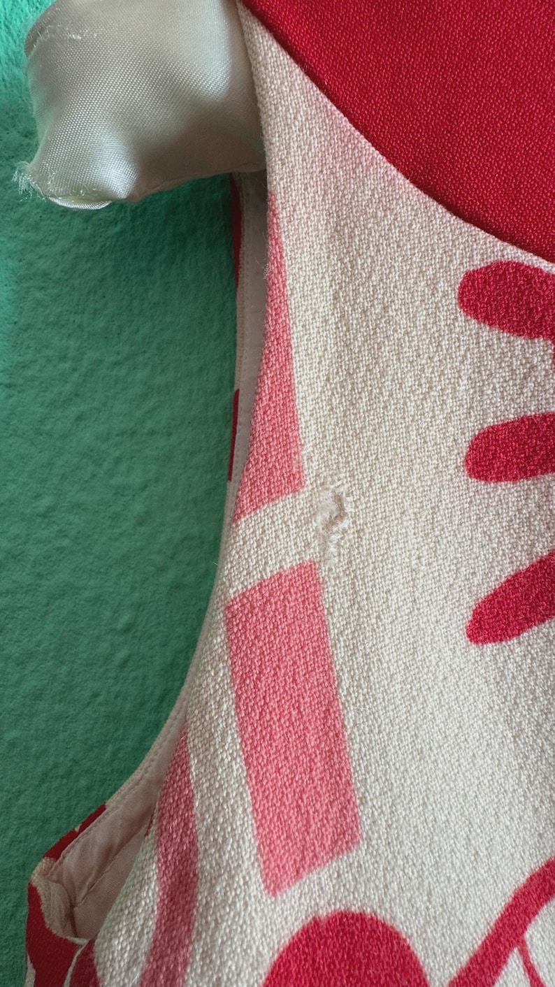 May include: A sleeveless garment featuring a textured off-white fabric with a bold red and pink geometric pattern. The design includes a solid red section at the top and a light pink lining visible along the armhole seam, showcasing a woven textile.