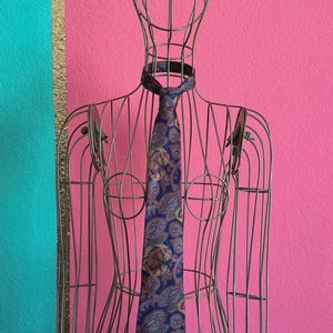 Könnte beinhalten: Eine blaue Krawatte mit einem mehrfarbigen Paisleuchtigen Paisley-Muster in Gold-, Rot- und Dunkelblautönen, präsentiert auf einer Draht-Schaufensterpuppe. Die Krawatte ist mit einem Standardknoten am Hals der Drahtstruktur gebunden und hängt gerade herab.