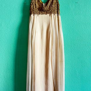 May include: A sleeveless floor-length dress featuring a cream-colored skirt with a sheer overlay and a bodice embellished with gold-toned embroidery, sequins, and a central floral accent at the V-neckline, hanging on a padded hanger.