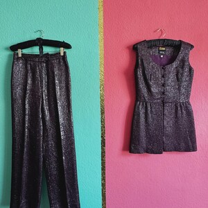 Könnte beinhalten: Ein zweiteiliges Outfit hängt vor einem farbenfrohen Hintergrund. Das Set umfasst eine Hose mit weitem Bein und ein ärmelloses Oberteil, beide aus einem dunklen, strukturierten Stoff mit dezentem Glanz. Das Oberteil hat eine Knopfleiste und einen Rundhalsausschnitt.