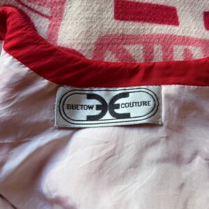 May include: A white garment interior featuring a red trim along the neckline and a rectangular fabric label sewn onto the lining. The label displays the text BUETOW COUTURE inside a stylized oval graphic. A pink and white patterned fabric is visible.