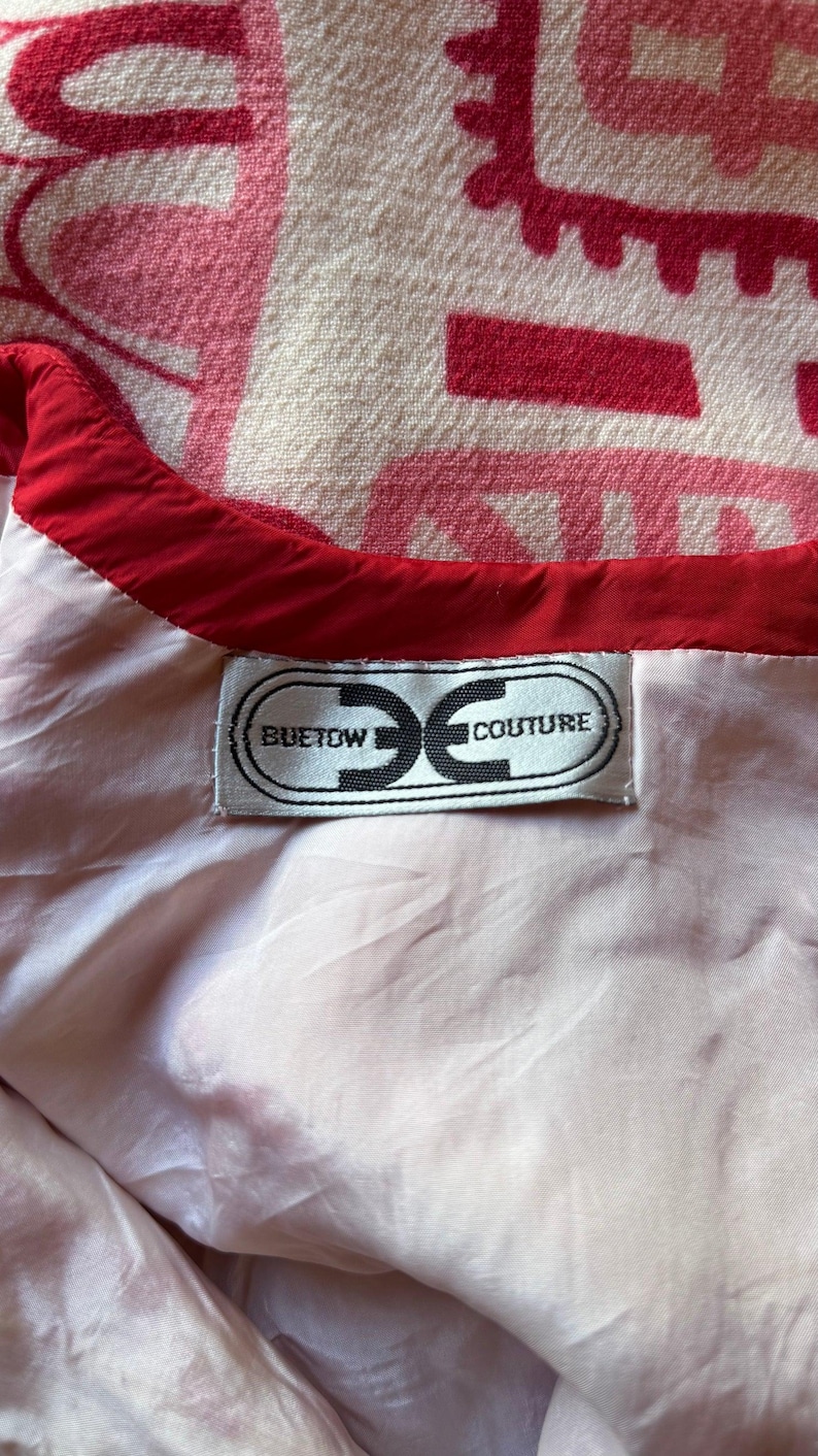 May include: A white garment interior featuring a red trim along the neckline and a rectangular fabric label sewn onto the lining. The label displays the text BUETOW COUTURE inside a stylized oval graphic. A pink and white patterned fabric is visible.