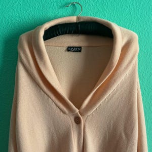 Può includere: Un cardigan color pesca chiaro con collo a scialle e chiusura con un solo bottone. Il maglione è appeso a una gruccia imbottita nera su uno sfondo turchese. Il nome del marchio "SANDINI" è visibile su un'etichetta.
