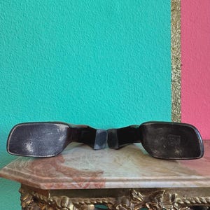 May include: Black vintage sunglasses with a rectangular frame. The sunglasses are resting on a marble-topped table with ornate gold detailing. The background features a teal and pink wall with a gold stripe.