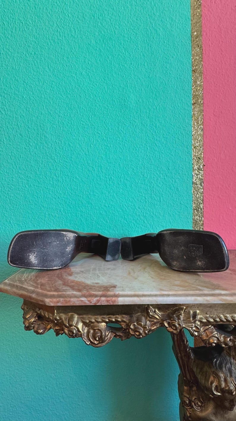 May include: Black vintage sunglasses with a rectangular frame. The sunglasses are resting on a marble-topped table with ornate gold detailing. The background features a teal and pink wall with a gold stripe.