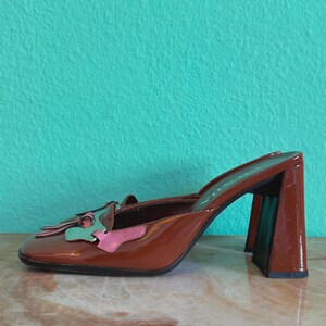 May include: Brown patent leather heeled mules with a square toe and block heel. The shoe features a decorative accent with pink and green details. The heel is approximately 3 inches high.
