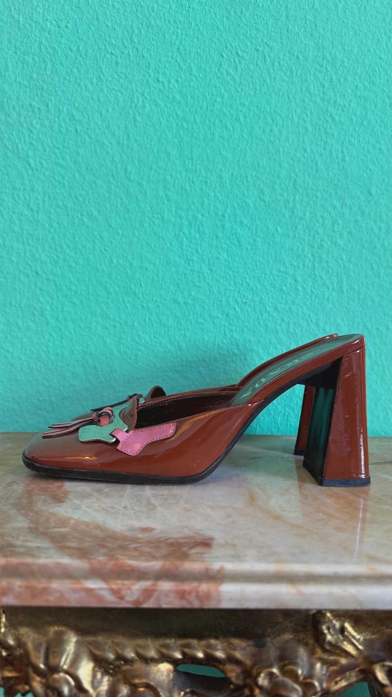 May include: Brown patent leather heeled mules with a square toe and block heel. The shoe features a decorative accent with pink and green details. The heel is approximately 3 inches high.