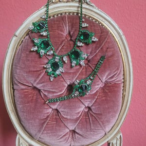 Può includere: Un set di collana e bracciale con strass verdi esposto su una sedia di velluto rosa. I gioielli presentano grandi pietre color smeraldo e strass trasparenti più piccoli. La sedia ha una cornice in legno verniciato oro.