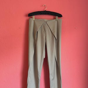 Può includere: Pantaloni beige caratterizzati da un design a vita alta con un distintivo dettaglio a pannello frontale sovrapposto. I pantaloni includono una chiusura centrale con bottone, una zip e gambe dritte, appesi a una gruccia in velluto nero.