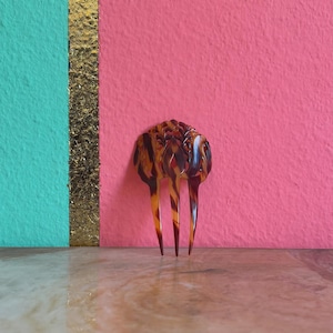 Può includere: Un pettine per capelli con motivo tartarugato marrone e nero, caratterizzato da una parte superiore decorativa intagliata e tre denti lunghi e sottili rivolti verso il basso. Il design è simmetrico, con intagli complessi nella parte superiore.