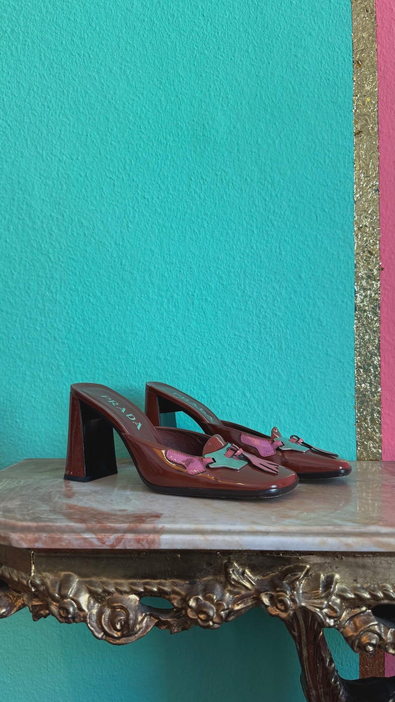 May include: A pair of brown leather Prada mules with block heels. The shoes feature a square toe and are adorned with pink and teal accents. The word "PRADA" is visible on the side of one shoe.