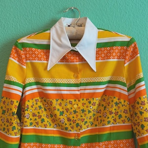 May include: A vintage button-down shirt with a white collar and long sleeves. The shirt features a vibrant pattern of horizontal stripes in yellow, orange, and green, with floral and geometric accents. The shirt is hanging on a hanger.