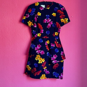 Puede incluir: Vestido azul marino con estampado floral en rojo, amarillo, azul y rosa. El vestido tiene mangas cortas y un detalle de peplum escalonado en la cintura. El vestido está colgado de una percha metálica.