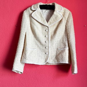 Può includere: Blazer color crema con tessuto strutturato, finiture bianche e quattro bottoni. La giacca ha due tasche anteriori ed è appesa a una gruccia nera su uno sfondo rosa acceso. Il colletto e i bordi del blazer sono delineati con finiture bianche.