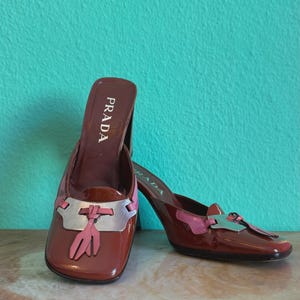 May include: A pair of Prada heeled mules in a glossy burgundy finish. The shoes feature a silver and pink accent on the vamp and a tassel detail. The Prada logo is visible on the insole.