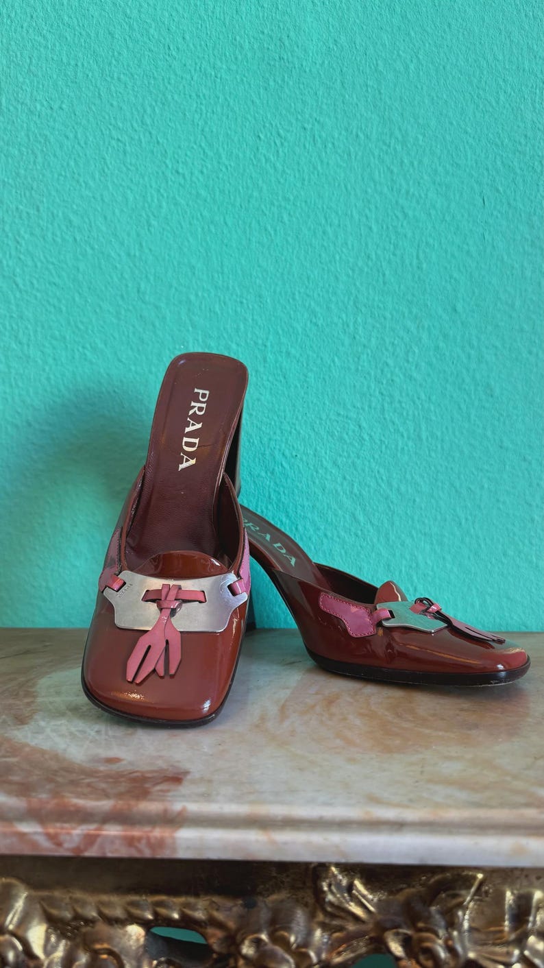 May include: A pair of Prada heeled mules in a glossy burgundy finish. The shoes feature a silver and pink accent on the vamp and a tassel detail. The Prada logo is visible on the insole.