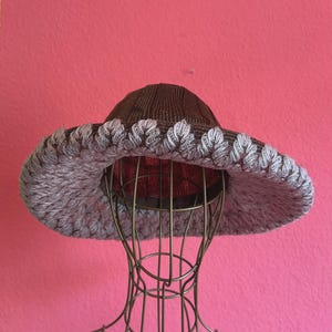 Può includere: Un cappello a tesa larga caratterizzato da una corona a coste marrone scuro e una tesa spessa e testurizzata in un materiale grigio chiaro simile al filato. La tesa presenta un motivo smerlato ripetitivo lungo il bordo, in netto contrasto.