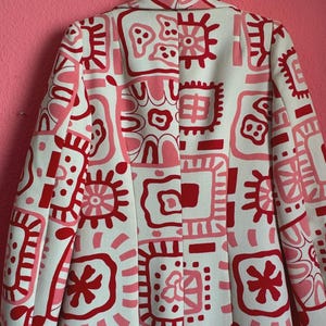 May include: A long-sleeved coat featuring a cream-colored base with a repeating pattern of abstract shapes, squares, and star-like motifs in shades of bright red and soft pink. The garment has a structured collar and a central vertical seam.