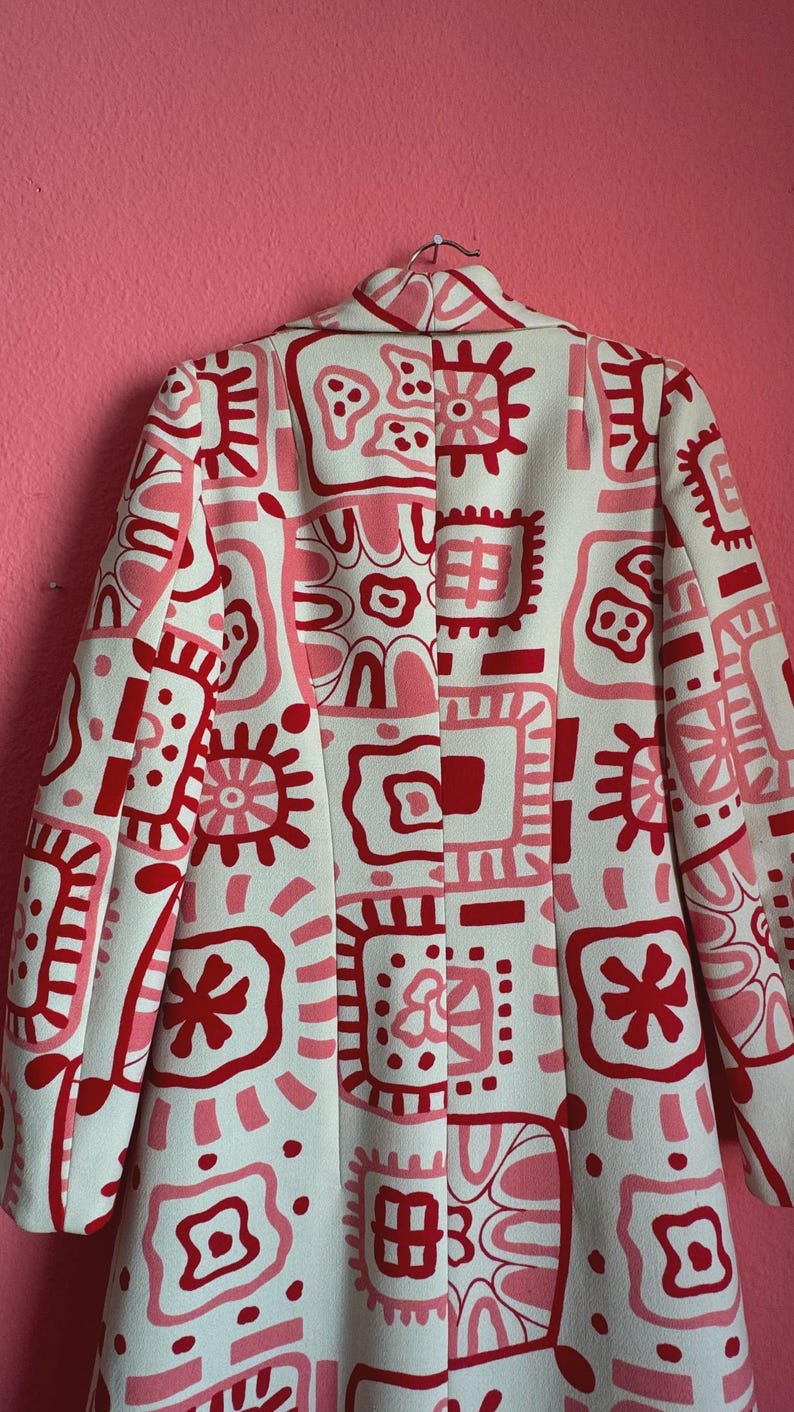May include: A long-sleeved coat featuring a cream-colored base with a repeating pattern of abstract shapes, squares, and star-like motifs in shades of bright red and soft pink. The garment has a structured collar and a central vertical seam.
