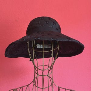 May include: A black wide-brimmed hat featuring a textured geometric pattern across the crown and brim. The hat includes a solid black ribbon band encircling the base of the crown and a white interior label with text visible inside the hat.