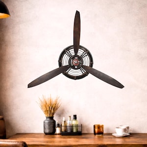 May include: A decorative wall clock designed to resemble an airplane propeller. The clock features three dark brown blades and a circular clock face with a black and bronze frame. The clock is mounted on a light-colored wall.