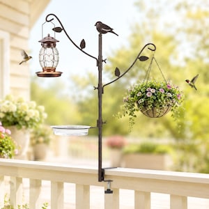 May include: A bronze-colored bird feeder and planter stand with a clamp for attaching to a deck railing. The stand features a hanging lantern-style bird feeder, a water dish, and a hanging basket filled with flowers. A decorative bird sits atop the stand.
