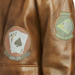 May include: Close-up of a brown leather jacket with two patches. One patch features playing cards, the other an eagle. The jacket has a pocket and is made of a distressed leather material.