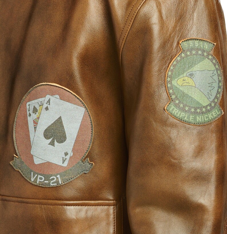 May include: Close-up of a brown leather jacket with two patches. One patch features playing cards, the other an eagle. The jacket has a pocket and is made of a distressed leather material.