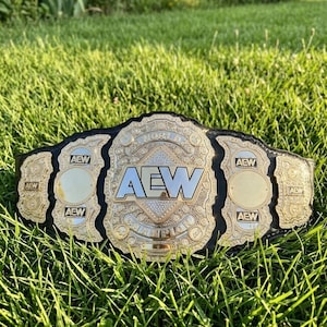 Könnte beinhalten: Ein gold-schwarzer Wrestling-Meisterschaftsgürtel mit den Buchstaben "AEW" in der Mitte. Der Gürtel hat goldene Akzente und ist vor einem Hintergrund aus grünem Gras platziert.
