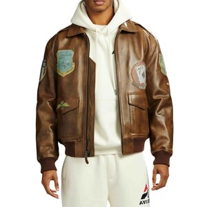 May include: A brown leather bomber jacket with multiple patches and pockets. The jacket has a zip-up front, a collar, and ribbed cuffs and hem. Patches include military-style emblems and card suits. The jacket is worn over a white hoodie.