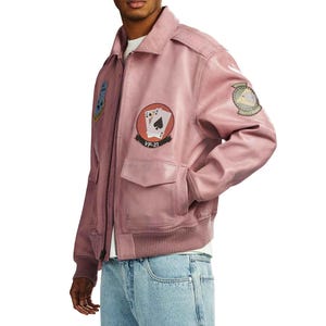 May include: A dusty rose leather jacket with a zip-up front, two front pockets, and a ribbed hem. The jacket features embroidered patches, including playing cards and a military emblem. The text "VP-21" is visible on one patch.