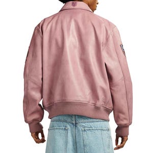 May include: A dusty rose leather jacket with a ribbed hem and cuffs. The jacket features a collar and a small emblem on the upper back. Patches are visible on the sleeves. The jacket is worn over light blue denim jeans.