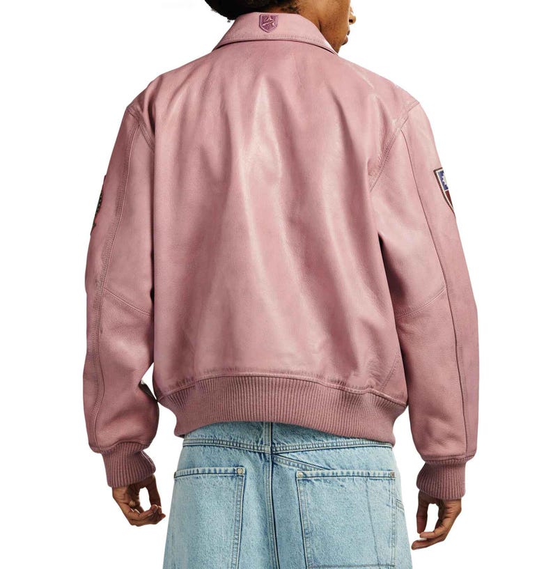 May include: A dusty rose leather jacket with a ribbed hem and cuffs. The jacket features a collar and a small emblem on the upper back. Patches are visible on the sleeves. The jacket is worn over light blue denim jeans.