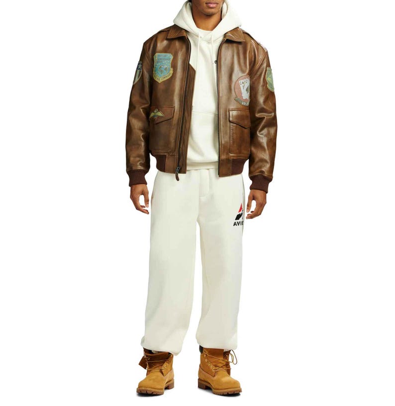 May include: A brown leather bomber jacket with patches on the sleeves and chest. The jacket has a zip-up front and two front pockets. The person is wearing a white hoodie, cream-colored sweatpants, and brown boots.