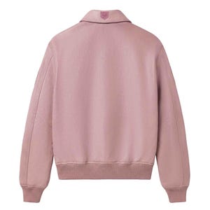 May include: A dusty rose bomber jacket with a collared neckline and ribbed cuffs and hem. The back of the jacket features a small emblem on the collar. The jacket has long sleeves and a relaxed fit, suitable for casual wear.