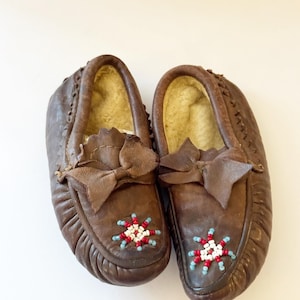 May include: A pair of brown leather moccasins with a bow detail and beaded floral design. The interior is lined with a light-colored material. The moccasins are a traditional style of footwear.