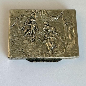 May include: A rectangular silver-toned box with a detailed relief scene. The relief depicts several figures in a landscape with foliage. The box has a hinged lid and a decorative edge, showcasing an antique or vintage style.