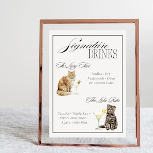 May include: A framed print with a rose gold border displays a "Signature DRINKS" menu. The menu features two cocktail recipes, "The Lucy Tini" and "The Lola Rita", with illustrations of cats and martini glasses. The ingredients are listed below each cocktail name.