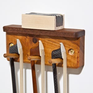 May include: A wooden violin bow holder with three slots, holding three bows with black and light-colored hair. A light-colored block of rosin sits on top. The holder is made of two types of wood, with brass screws.