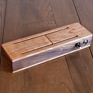 May include: A rectangular wooden audio device featuring a light-toned top surface with two horizontal slots and a dark-toned side panel. The side includes a single black rotary control knob and a circular metal input jack port for connectivity.