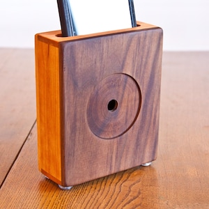 May include: A rectangular wooden phone stand crafted from dark walnut and lighter cherry wood. The front face features a circular recessed detail with a central hole, and the top has a slot for a mobile device. The stand rests on small clear feet.