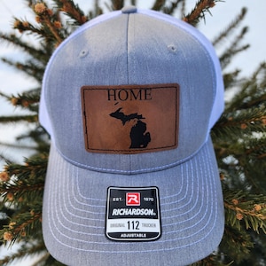 May include: A gray and white trucker hat with a leather patch featuring the word "HOME" above a black outline of the state of Michigan. The hat has a curved brim and an adjustable strap. The Richardson brand logo is visible.