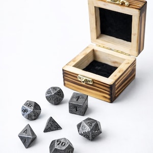 May include: A wooden box with a hinged lid and a black felt interior, accompanied by a set of seven metal dice. The dice are various geometric shapes, including a cube, a d20, and a d100.