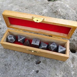 May include: A wooden box with a red velvet lining contains seven metal dice. The dice are grey with black etched numbers and patterns. The box is open, showing the dice, and has a gold-coloured clasp.