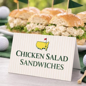 May include: Chicken salad sandwiches with green flags, served with golf balls and a sign that reads "CHICKEN SALAD SANDWICHES". The sign features a yellow map with a red flag.
