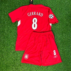 May include: A red soccer uniform set, including a jersey and shorts, with white accents. The jersey features the name "GERRARD" and the number "8" in white, along with a soccer ball graphic. The shorts also display the number "8".