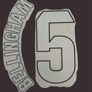 May include: White heat transfer vinyl with the name "BELLINGHAM" in a curved font and the number "5". The number has a dotted pattern on the top section and a black outline. The background is a dark grey.