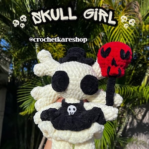 May include: A crocheted doll with a cream-colored body, black circular eyes, and a black dress featuring a small white skull emblem. The doll wears a black hat and holds a red skull-shaped accessory on a black stick. Text reads SKULL GIRL @crochetkareshop.