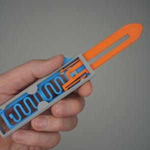 Könnte beinhalten: Ein 3D-gedrucktes, abstraktes Objekt, das in einer Hand gehalten wird. Das Objekt hat einen grauen, rechteckigen Rahmen mit blauen und orangefarbenen Innenteilen. Der orangefarbene Abschnitt hat ein gabelförmiges Design. Der blaue Abschnitt hat eine wellenförmige, rohrartige Struktur.