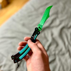 Puede incluir: Una navaja tipo mariposa con una hoja de color verde brillante con superficie texturizada y mangos negros. Los mangos tienen secciones recortadas que revelan una capa interior verde brillante. La navaja está abierta y tiene un cordón negro.